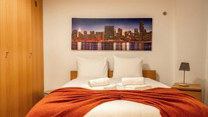 A hotel room with a bed, a cityscape painting, a wooden wardrobe, and a nightstand with a lamp. - Carob Apartment, Baixa, Lisbon (Lisboa)
