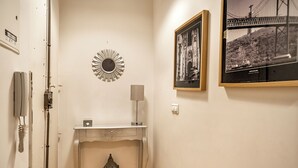 A hallway with a white wall, a framed photograph, a small table with a lamp, and a decorative mirror. - Carob Apartment, Baixa, Lisbon (Lisboa)