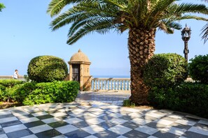 A checkered pathway leading to a building with a dome, surrounded by greenery and a view of the sea.