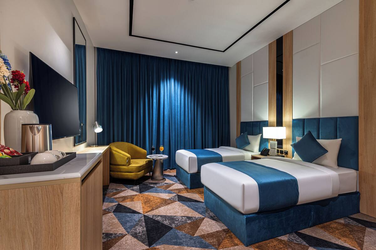A modern hotel room with a large bed, two bedside tables, a desk, a yellow armchair, and a patterned carpet.