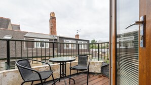 En balkon med to stole og et bord, udsigt over et boligområde med huse og skorstene.