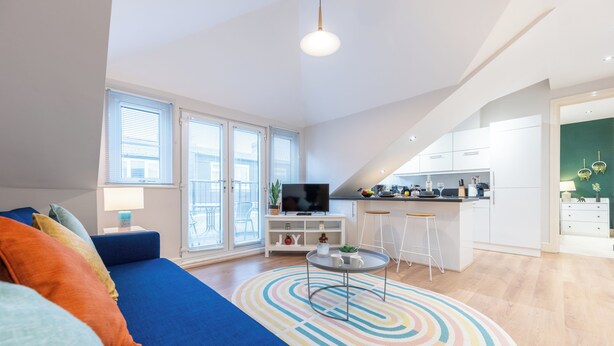 En moderne stue med en blå sofa, et fjernsyn og et køkkenområde med hvide skabe og en barstol.