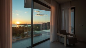 Ein modernes Zimmer mit einem großen Glasfenster, einer Bank und Blick auf einen Sonnenuntergang über dem Wasser.