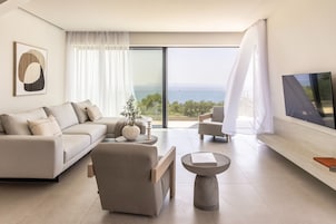 Una sala de estar moderna con un sofá, un sillón, una mesa de centro, un televisor de pantalla plana y un ventanal con vista al mar.