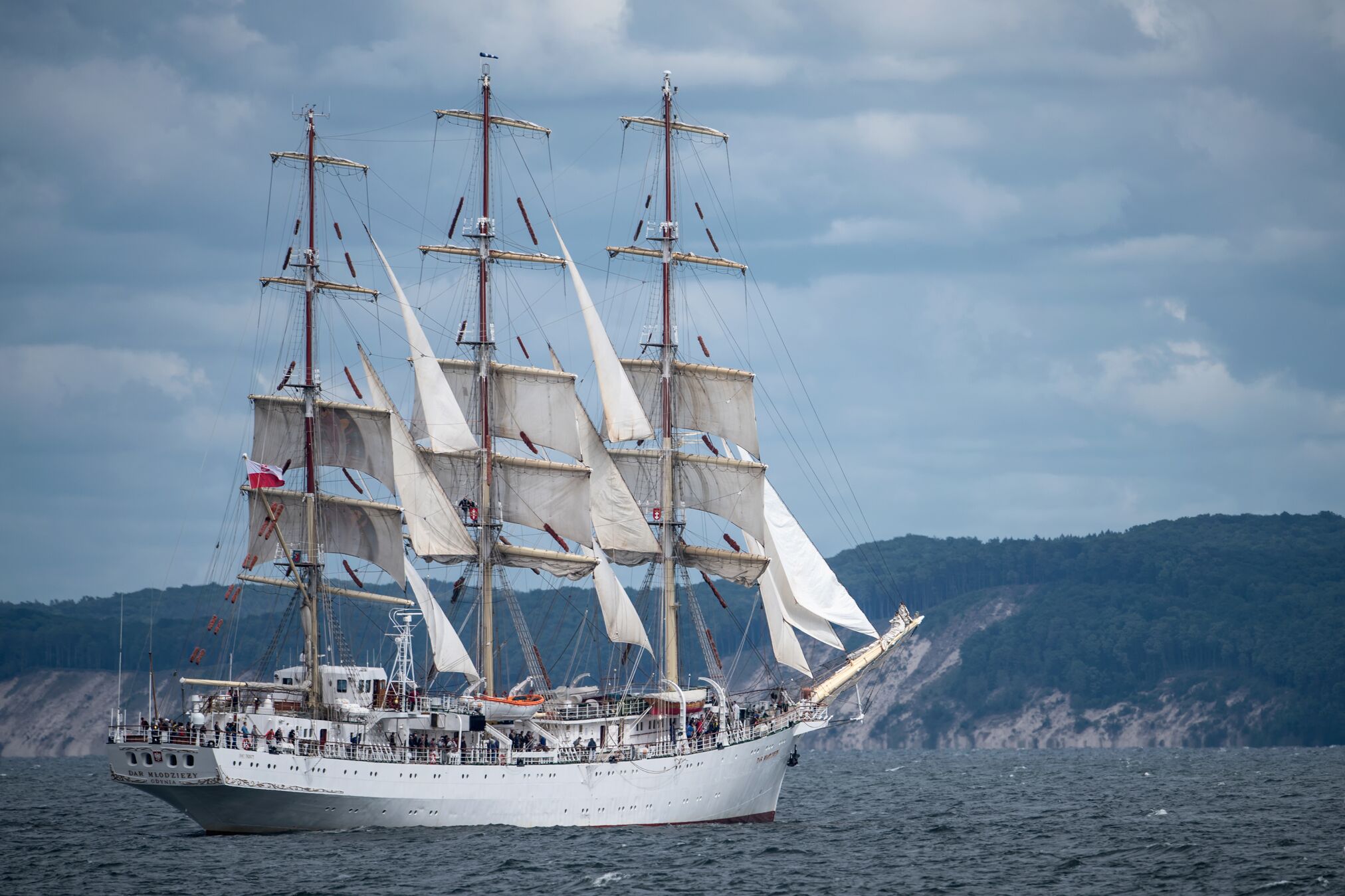 A large sailing ship with multiple masts and sails, docked at a harbor with a forested coastline in the background.
