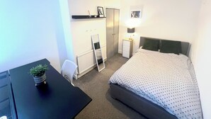 Una habitación de hotel con cama, escritorio, silla, radiador, armario y una planta pequeña.