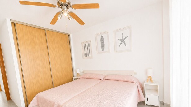 A bedroom with a bed, a ceiling fan, a wardrobe, a nightstand, and wall art. - Apartamento Las Tres Piedras Conil (conil de la frontera)