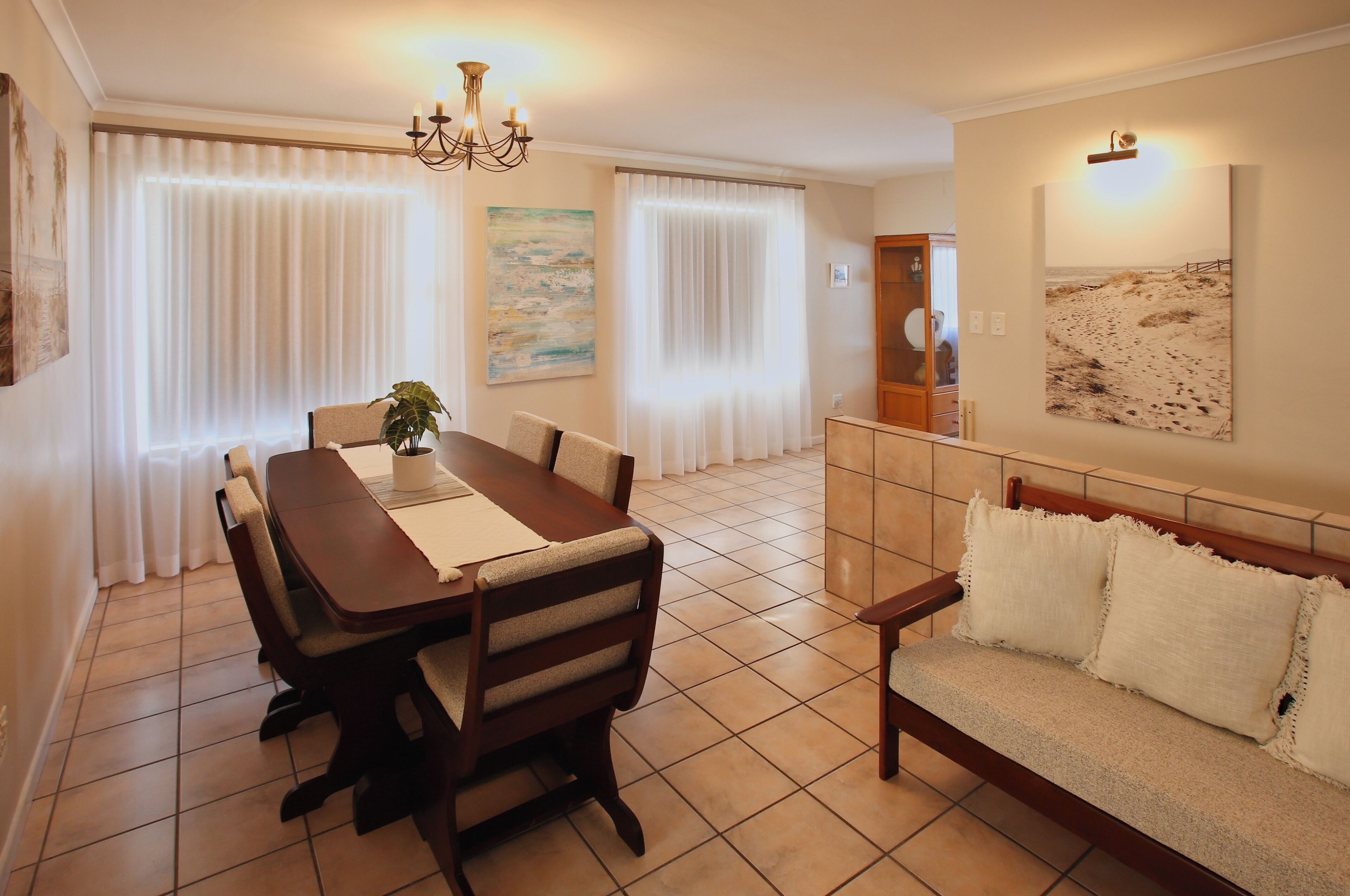 A dining area with a wooden table and chairs, a sofa with cushions, a chandelier, and a painting on the wall.