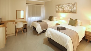 Ein Hotelzimmer mit zwei Betten, einem Schreibtisch, einem Stuhl, einem Mülleimer und einem Bild an der Wand.