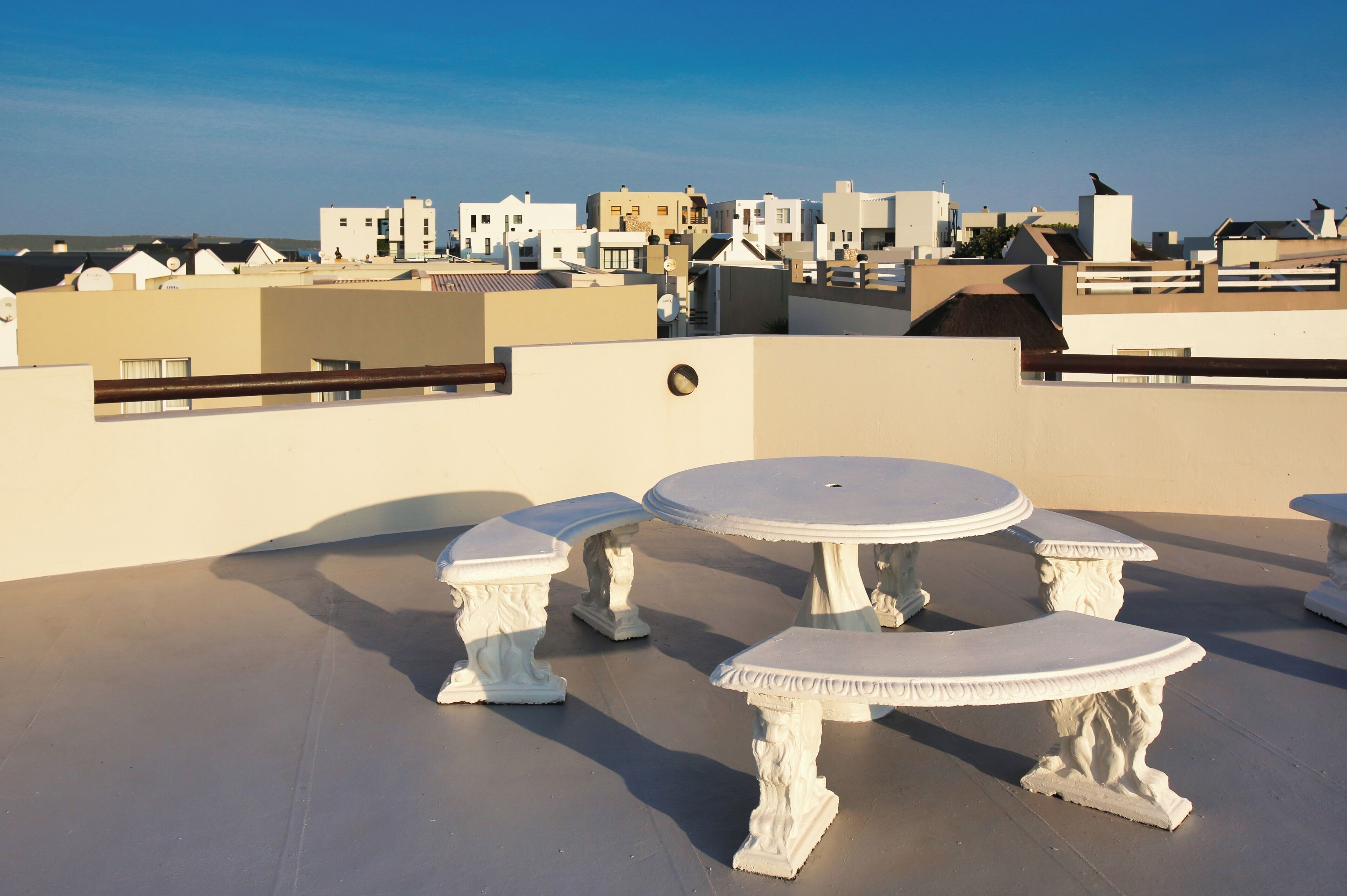 A rooftop terrace with a round table and two benches, surrounded by a low wall and a railing.