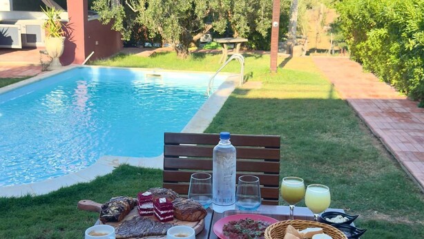 A poolside breakfast setup with a variety of foods and drinks, a wooden table, and a pool in the background. - La Cascina (Sidi Bou Ali)