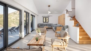 A spacious living area with a wooden table, chairs, and a sofa. There is a staircase with a cat decoration and a view of the outdoors through large windows. - Kourtaliotis Villa Crete (Agios Vasileios)