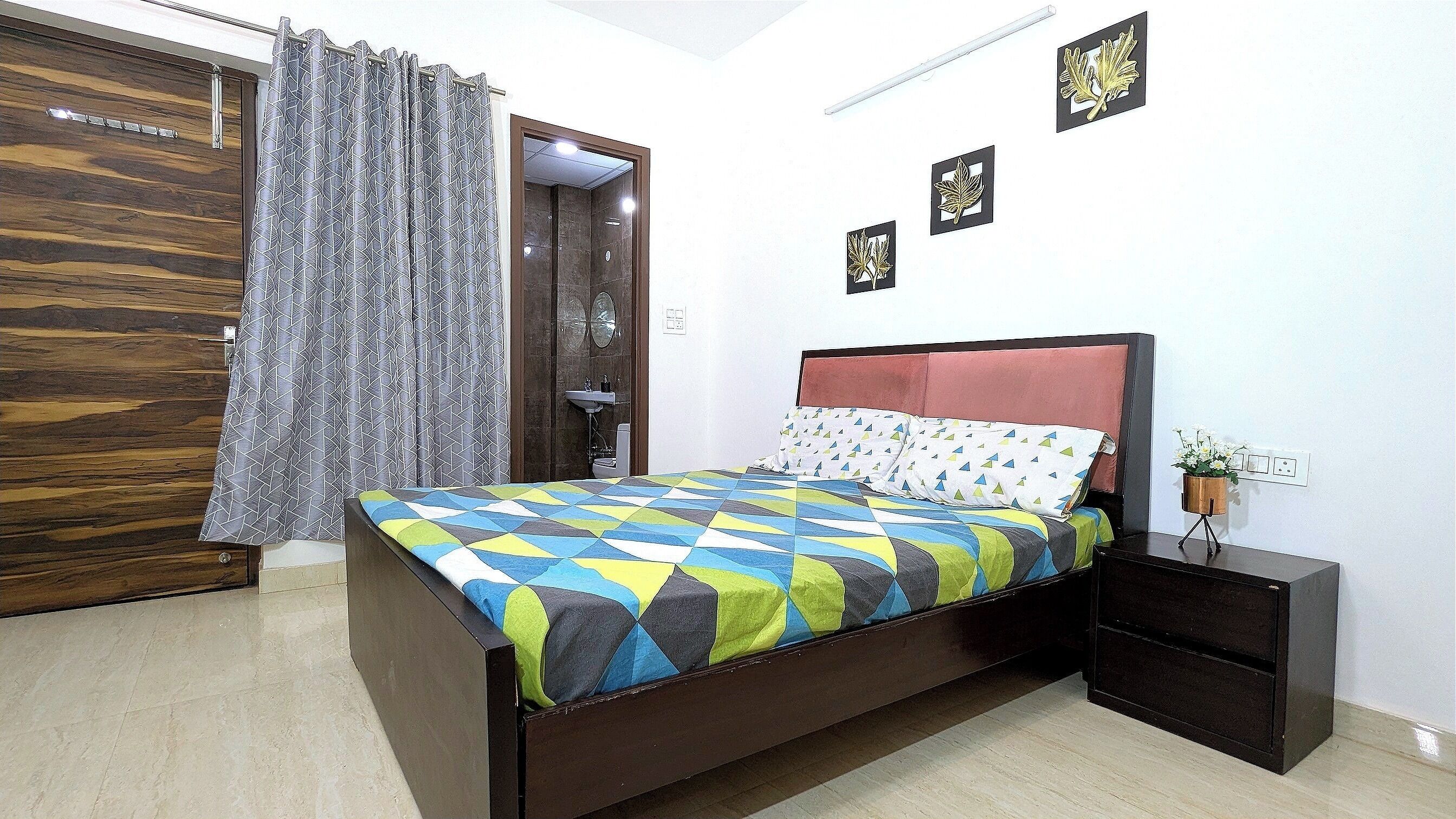 A bedroom with a wooden wardrobe, a bed with a colorful geometric-patterned bedspread, a bedside table with a plant, and a wall-mounted light fixture.