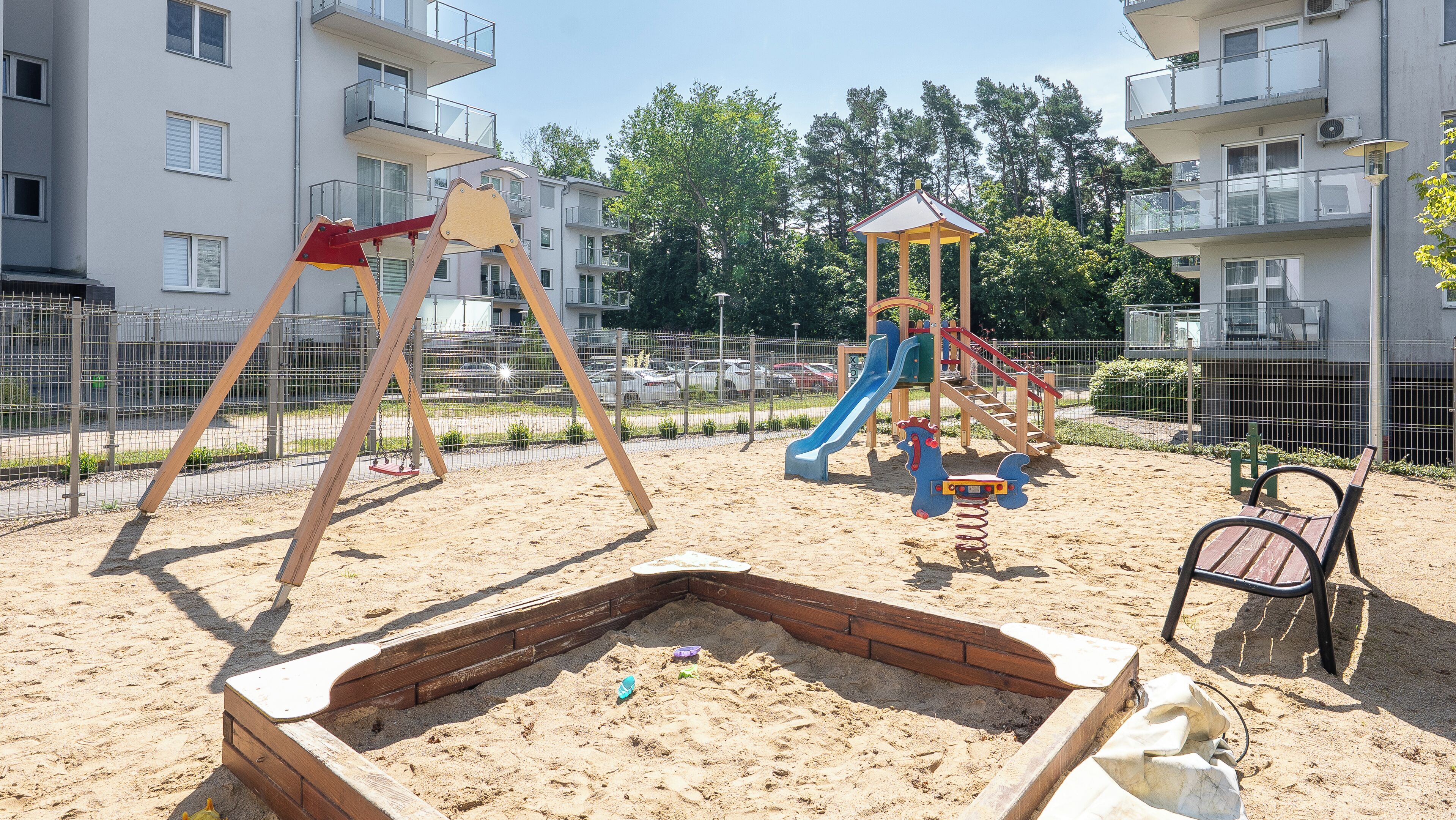 A playground with swings and a slide, surrounded by a fence and residential buildings.