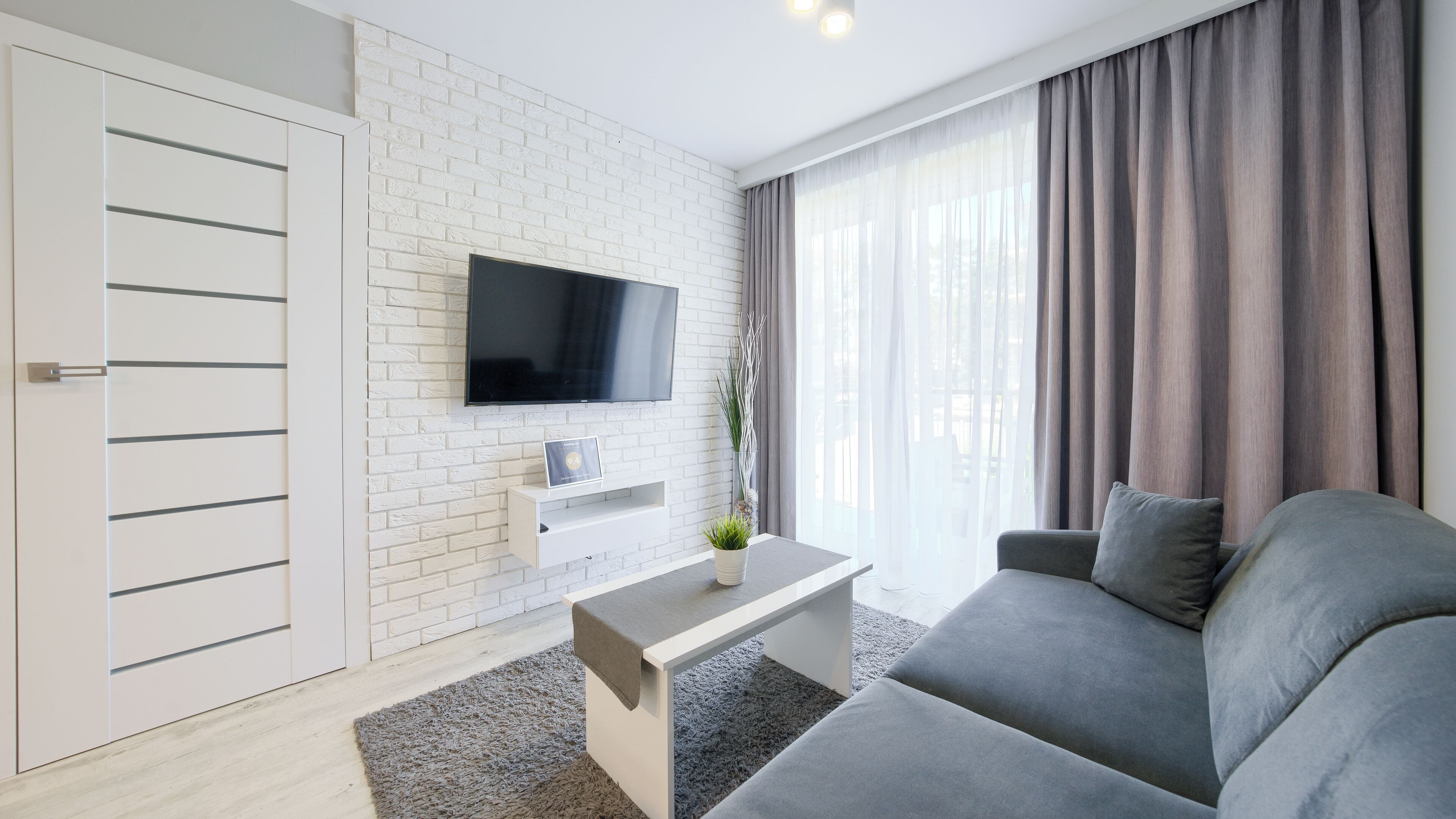 A modern living room with a white brick wall, a flat-screen TV mounted above a white console table, a grey sofa, and a grey rug.