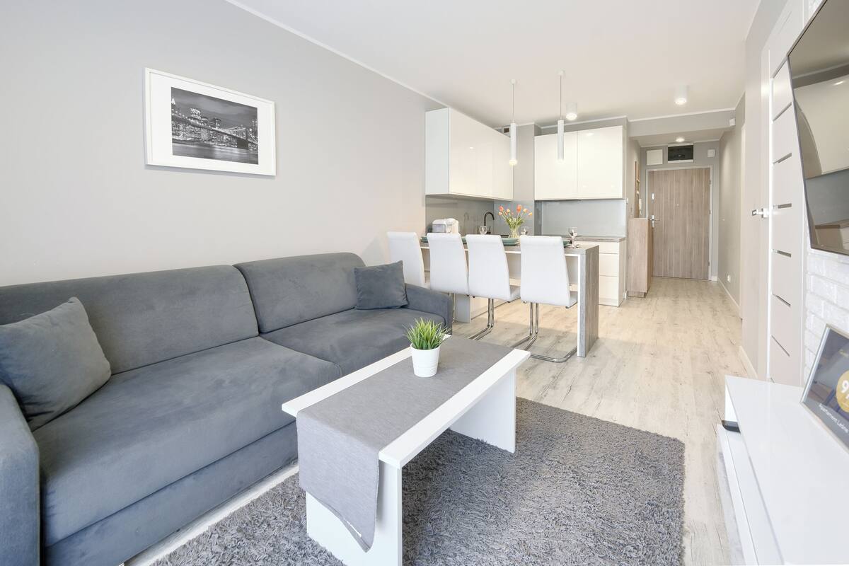 A modern living room with a grey sofa, a small white table with a plant, and a grey rug. There is a dining area with white chairs and a kitchen in the background.