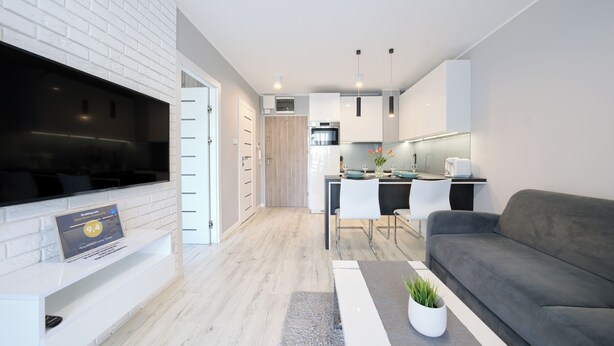 A modern living room with a grey sofa, a flat-screen TV mounted on a white brick wall, a small dining area with a table and chairs, and a kitchen area with white cabinets and a built-in oven. - Apartamenty Sun & Snow Leśne Tarasy (Dziwnówek)