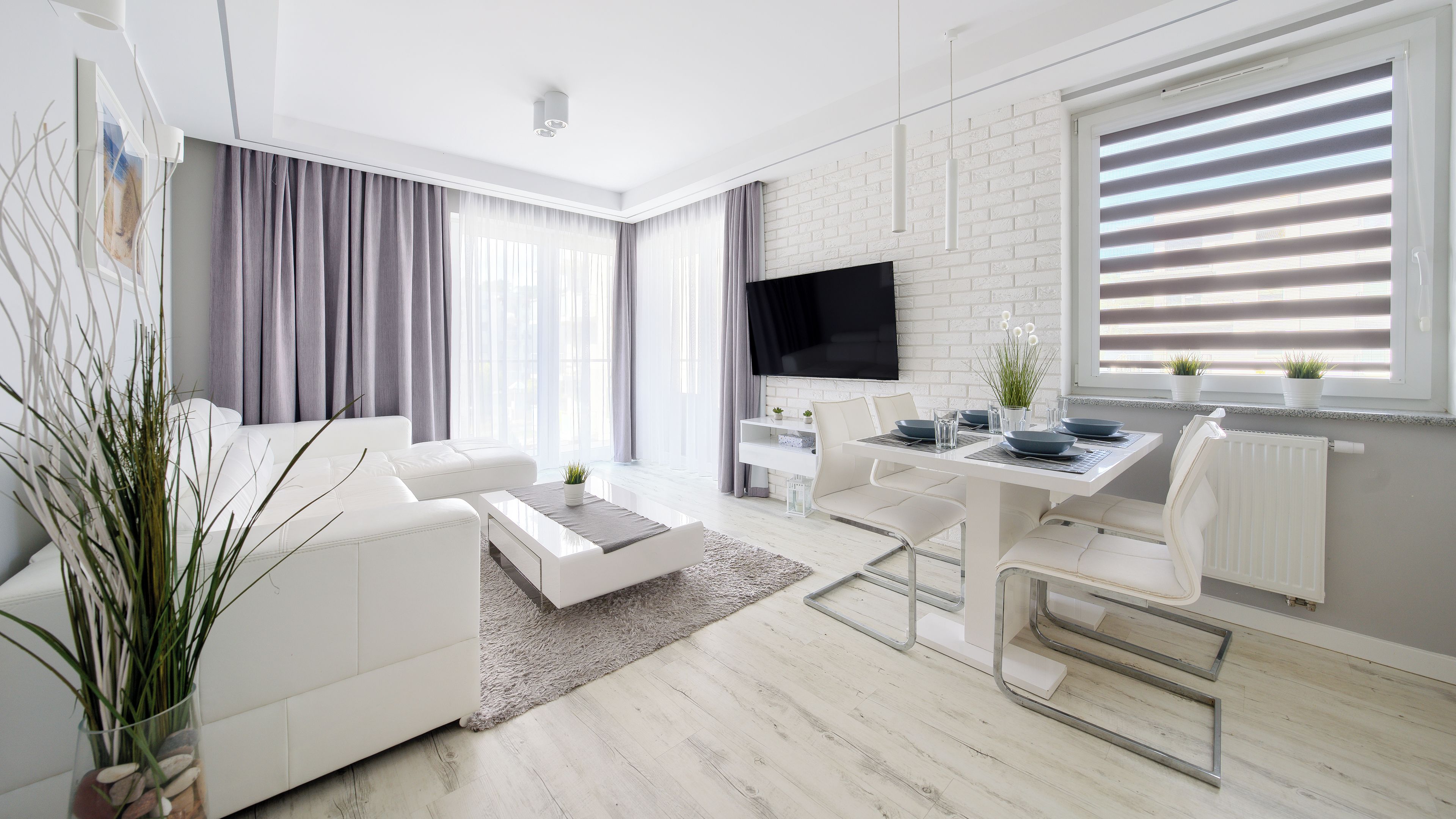 A modern living room with a white sofa, a dining table with chairs, a flat-screen TV mounted on a white brick wall, and a large window with blinds.