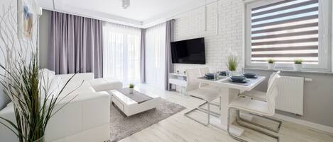 A modern living room with a white sofa, a dining table with chairs, a flat-screen TV mounted on a white brick wall, and a large window with blinds.
