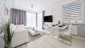 A modern living room with a white sofa, a dining table with chairs, a flat-screen TV mounted on a white brick wall, and a large window with blinds.