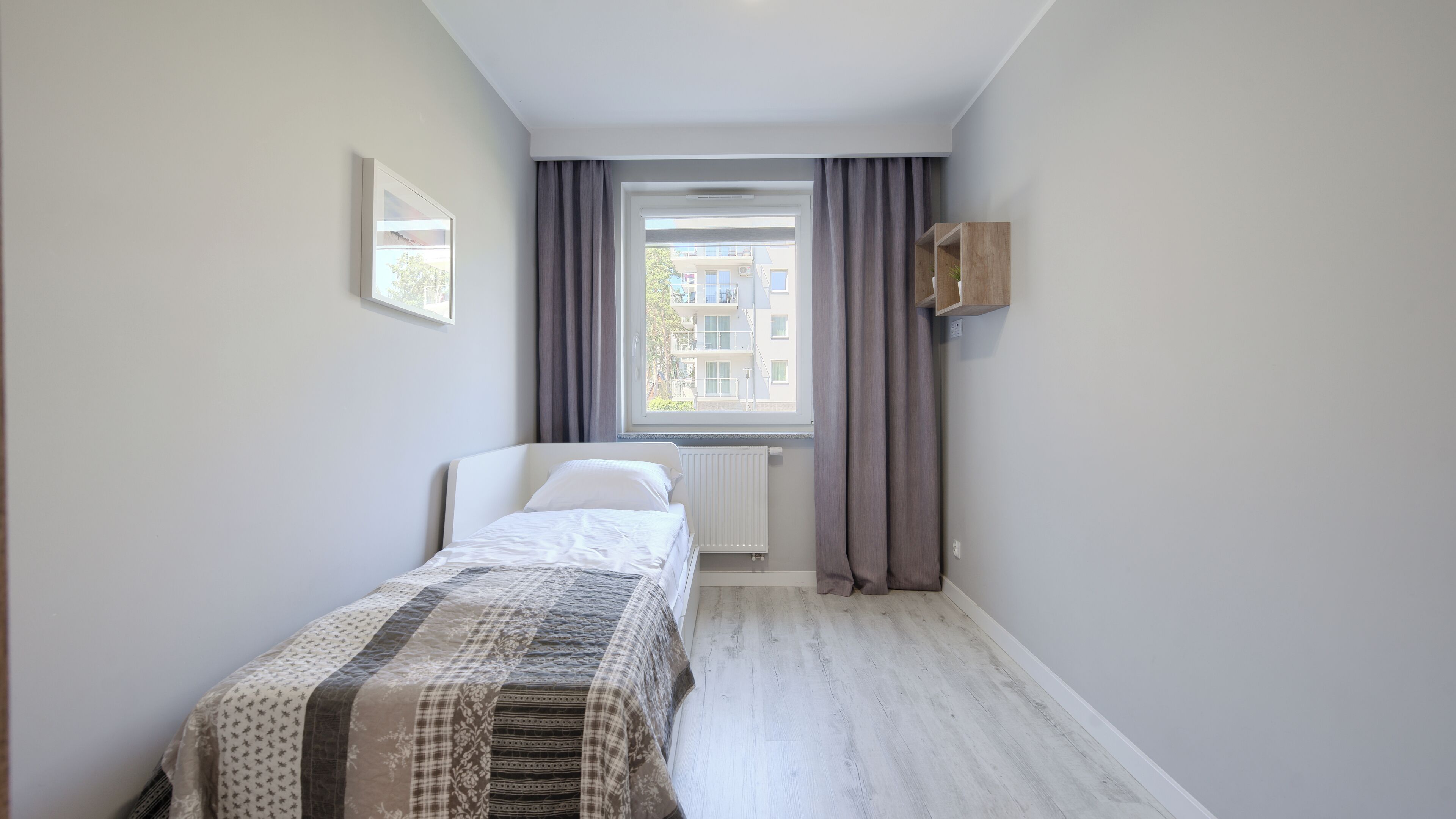 A small, neatly arranged bedroom with a single bed, a window with curtains, a radiator, and a small wooden shelf.