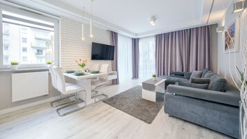 Una sala de estar moderna con un sofá gris, una mesa de comedor con sillas blancas, un televisor montado en la pared y ventanas grandes con cortinas.