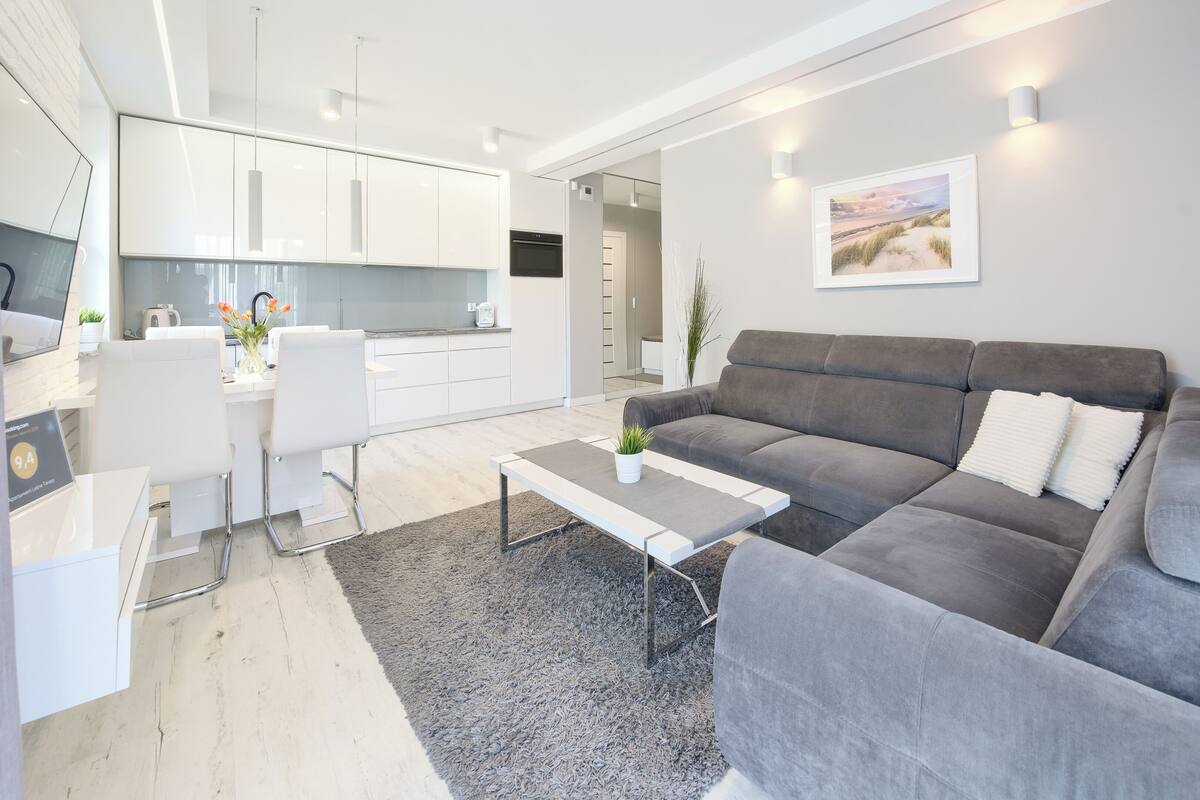 A modern living room with a grey sofa, a coffee table, and a dining area with white cabinets and a kitchen in the background.