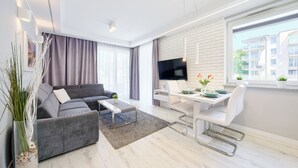 A modern living room with a grey sectional sofa, a coffee table, a dining area with a white table and chairs, a flat-screen TV mounted on the wall, and a large window with a view of buildings. - Apartamenty Sun & Snow Leśne Tarasy (Dziwnówek)