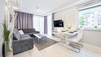 Una sala moderna con un sofá seccional gris, una mesa de centro, un comedor con una mesa y sillas blancas, un televisor de pantalla plana montado en la pared y un ventanal con vistas a edificios.
