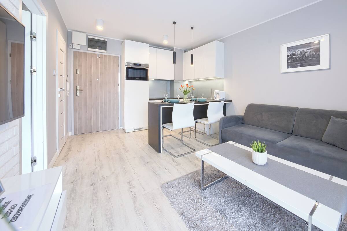 A modern apartment with a grey sofa, a dining area with white chairs, and a kitchen with white cabinets.