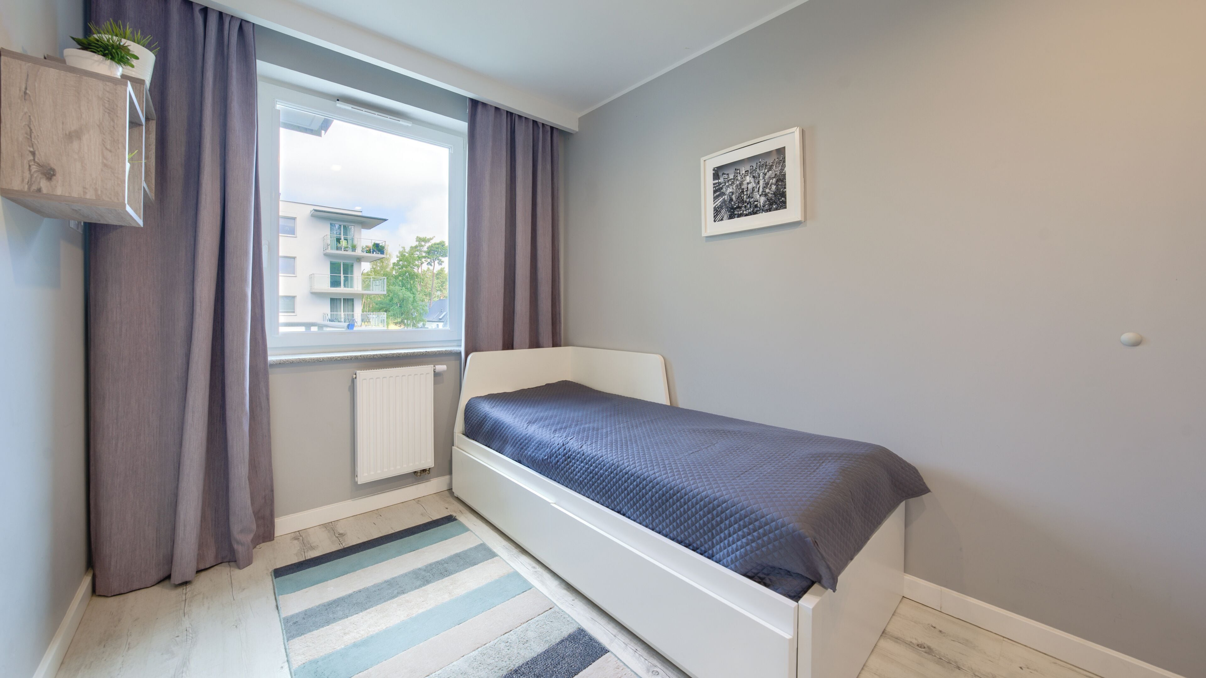 A bedroom with a bed, a window with curtains, a radiator, a striped rug, and a framed picture on the wall.