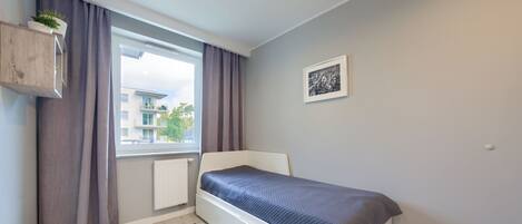 Quarto com cama, janela com cortinas, radiador, tapete listrado e um quadro na parede.