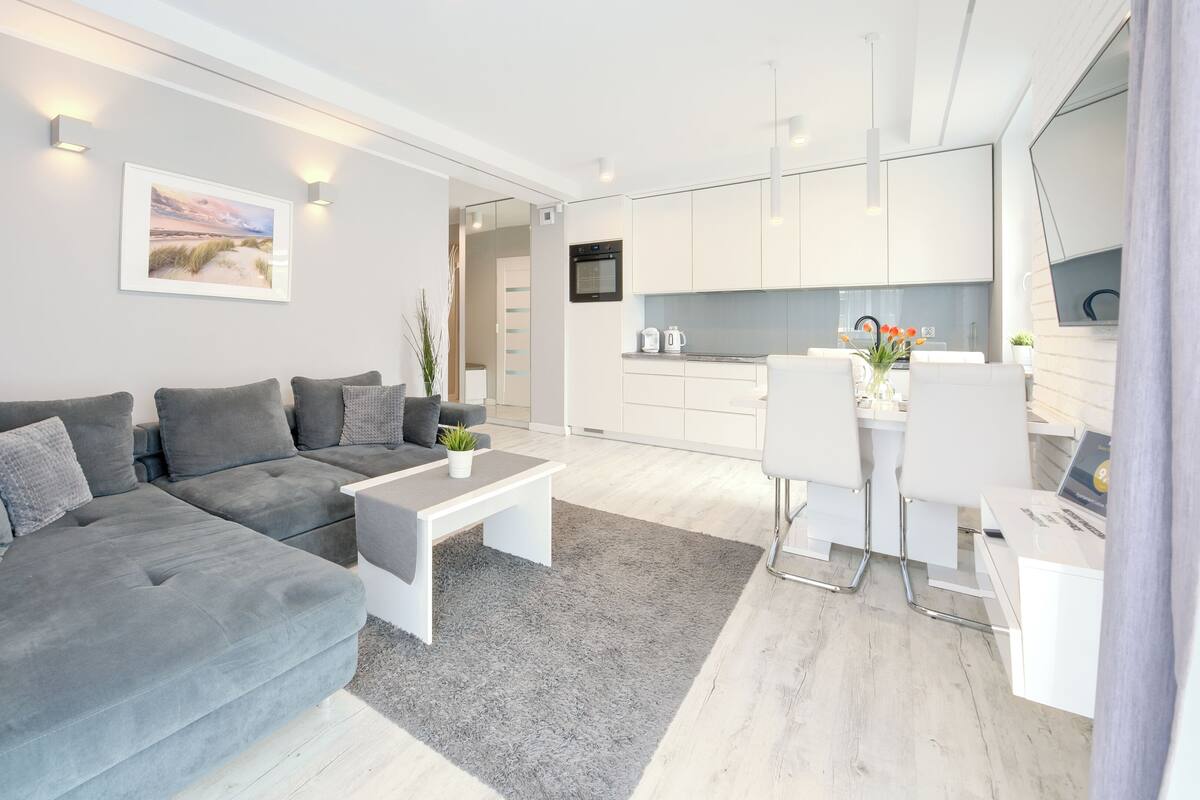 A modern living room with a grey sectional sofa, a white coffee table, and a kitchen area with white cabinets and a flat-screen TV.