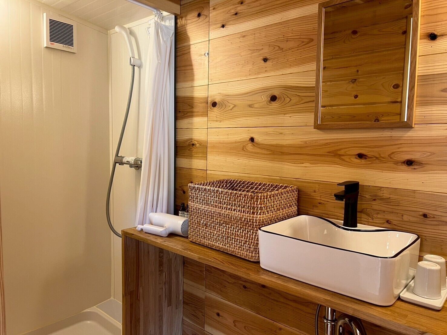 A bathroom with a wooden wall, a white sink with a black faucet, a woven basket, and a shower with a white curtain.