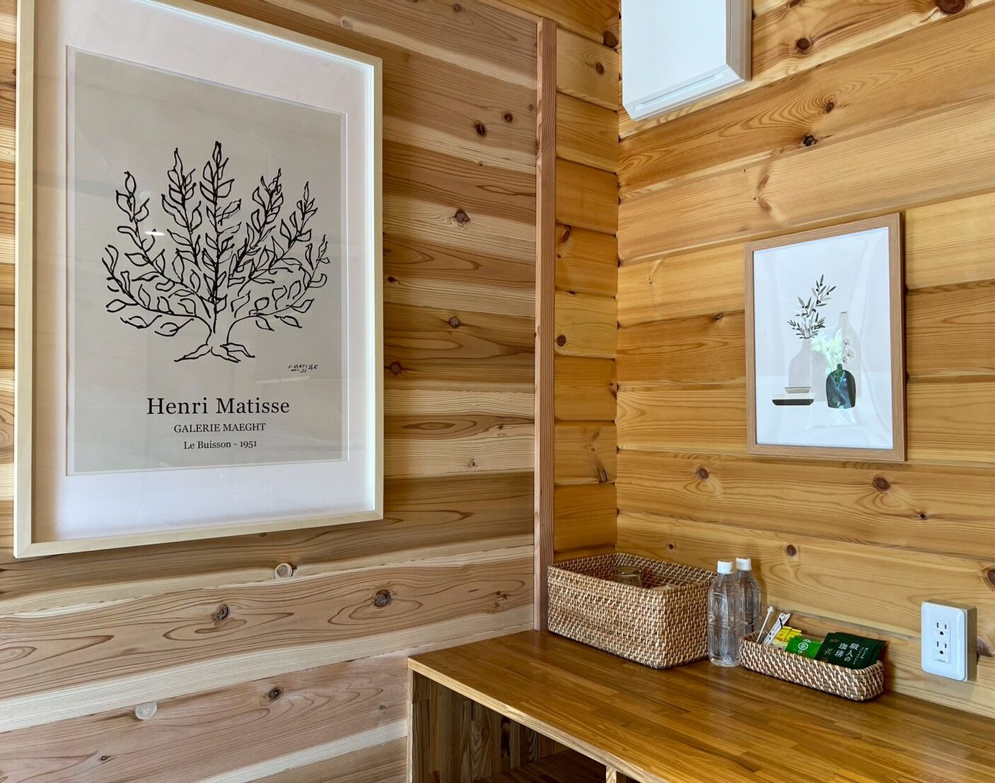 Una pared de madera con dos cuadros enmarcados, una cesta tejida y una mesita con botellas y un recipiente.