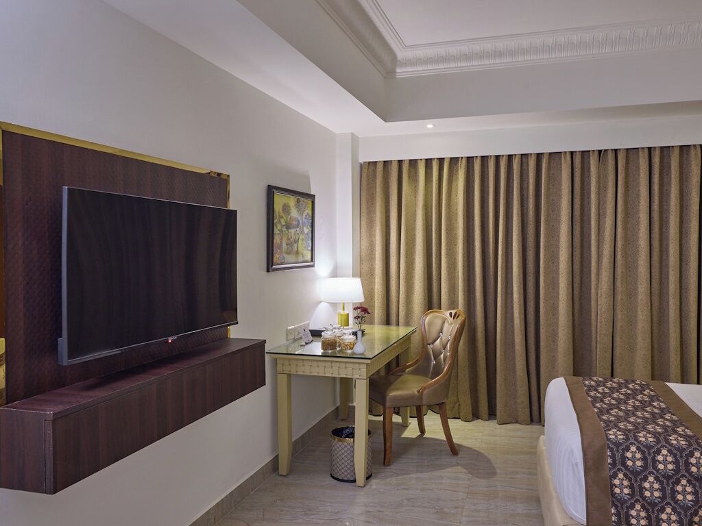 A hotel room with a flat-screen TV, a desk with a lamp, a chair, a bed, and a large window with curtains.