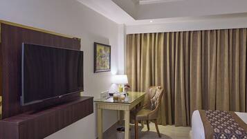 A hotel room with a flat-screen TV, a desk with a lamp, a chair, a bed, and a large window with curtains.