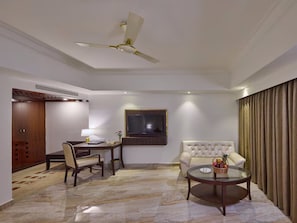 A hotel room with a dining area, a sofa, a TV mounted on the wall, and a ceiling fan.