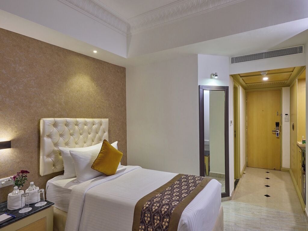 Habitación de hotel con una cama grande, una mesita de noche, un espejo, una puerta y un pasillo que lleva a un baño.