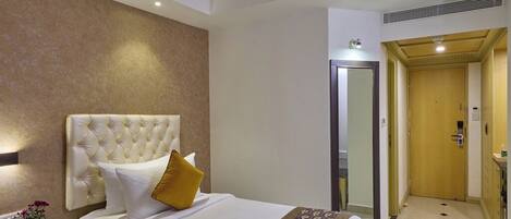 Habitación de hotel con una cama grande, una mesita de noche, un espejo, una puerta y un pasillo que lleva a un baño.