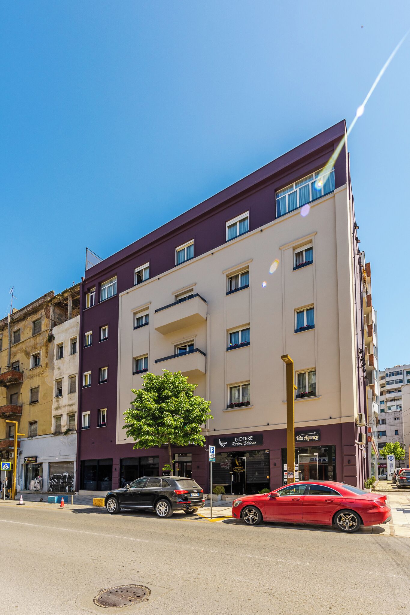 Un edificio de varios pisos con fachada plana, varias ventanas y un árbol en primer plano.