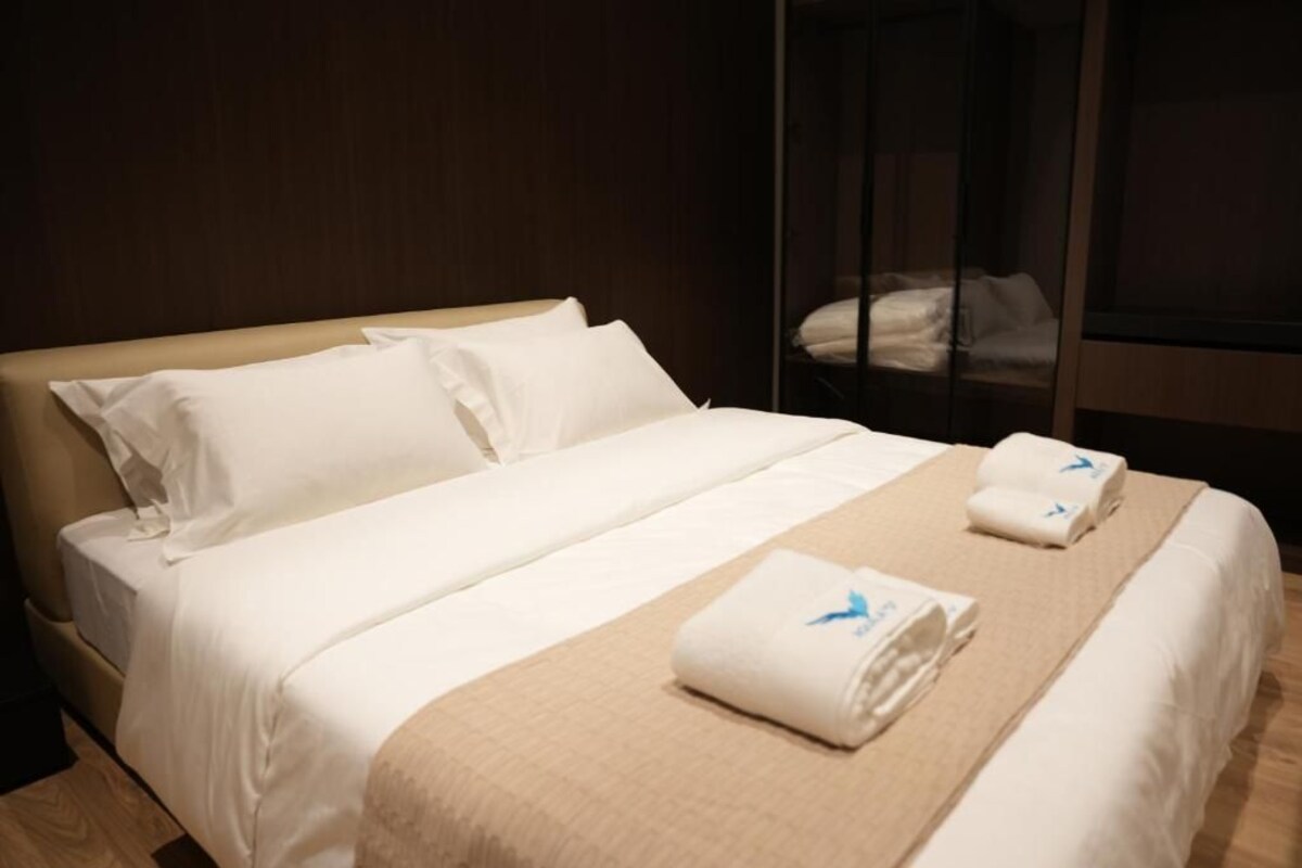 Una cama bien tendida con sábanas blancas y una manta color beige, suelo de madera y una ventana con persianas.