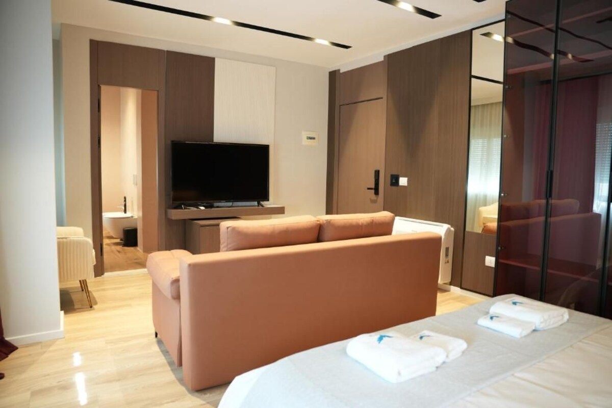 A modern hotel room with a large bed, a sofa, a flat-screen TV, and a glass partition.