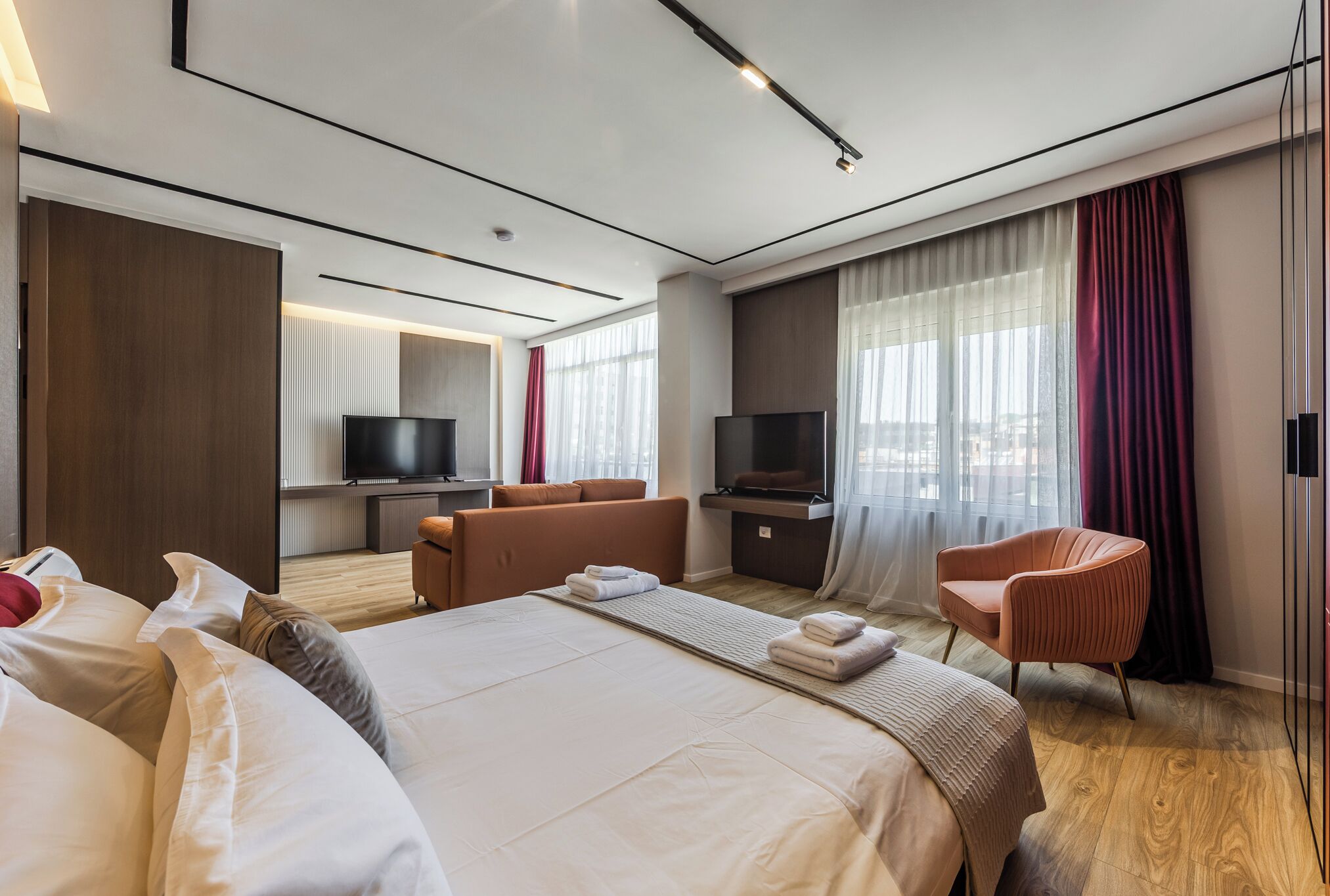 A modern hotel room with a large bed, a seating area, a TV, and a view of the city.
