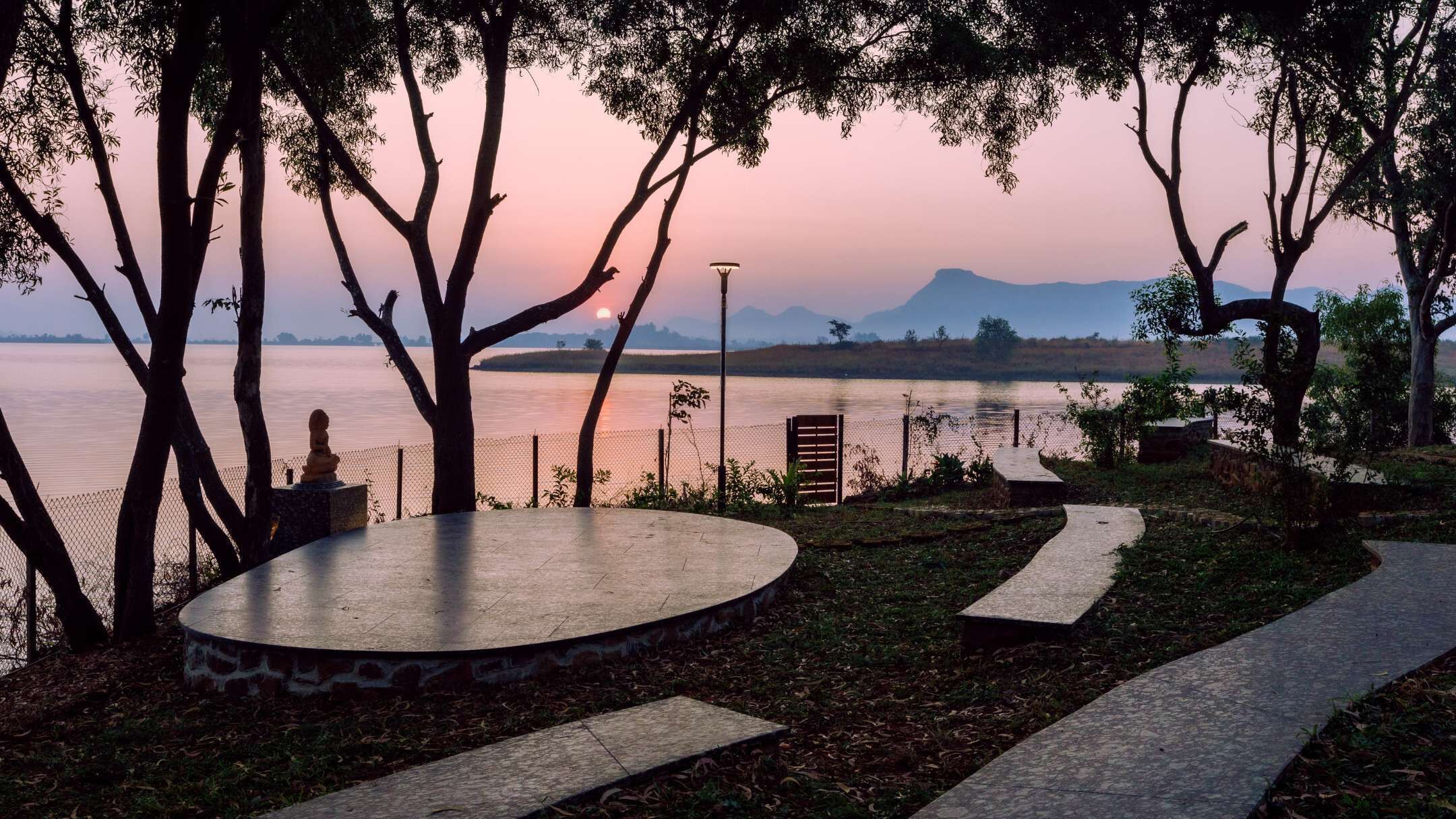 A circular stone seating area with a statue, surrounded by trees and a view of a calm lake with mountains in the distance.