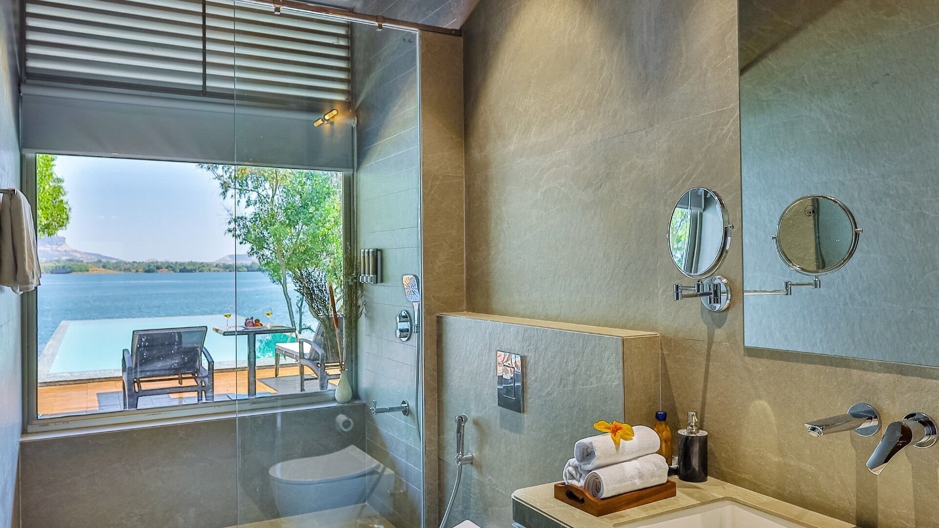 Un baño moderno con cabina de ducha de vidrio, lavamanos con espejo y vistas a un lago y una zona de estar al aire libre.
