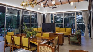 A spacious dining area with a wooden table and chairs, a sofa, and a television. Large windows provide a view of the outdoors. - Nature's Island (Trimbak)