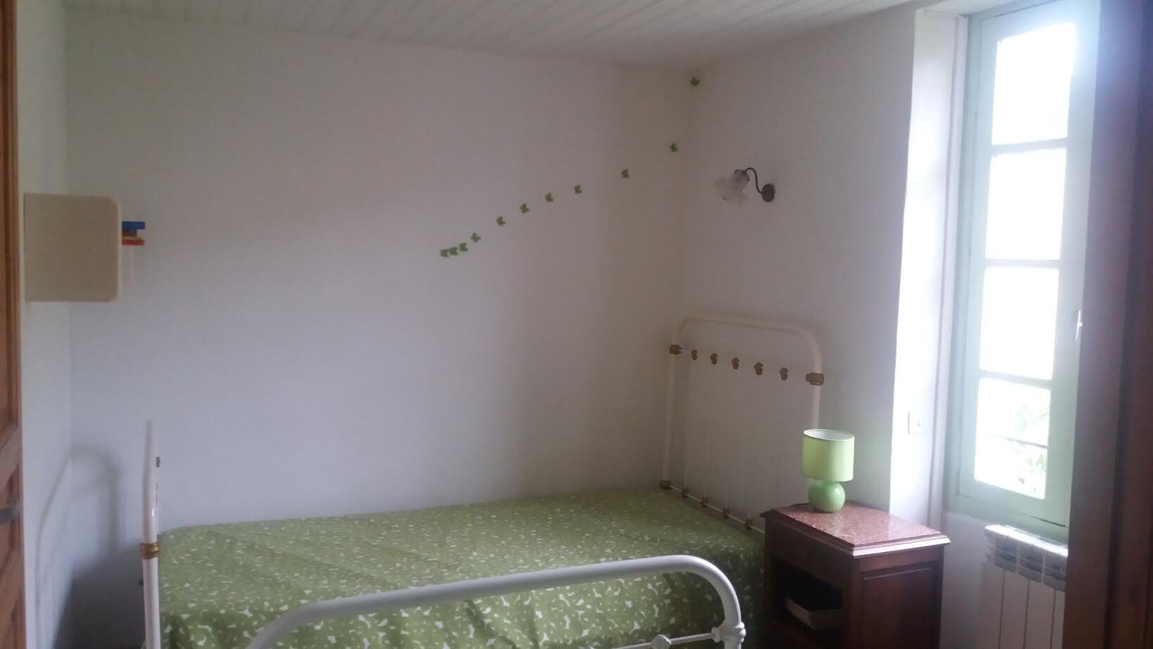 A small, single-bed room with a green bedspread, a bedside table with a lamp, and a window with a view of greenery.