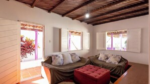 A room with a wooden ceiling, two sofas with floral cushions, a red ottoman, and a view through a door with shutters. - Casa Pet-friendly com Piscina de Frente Para o mar (Ilhéus)