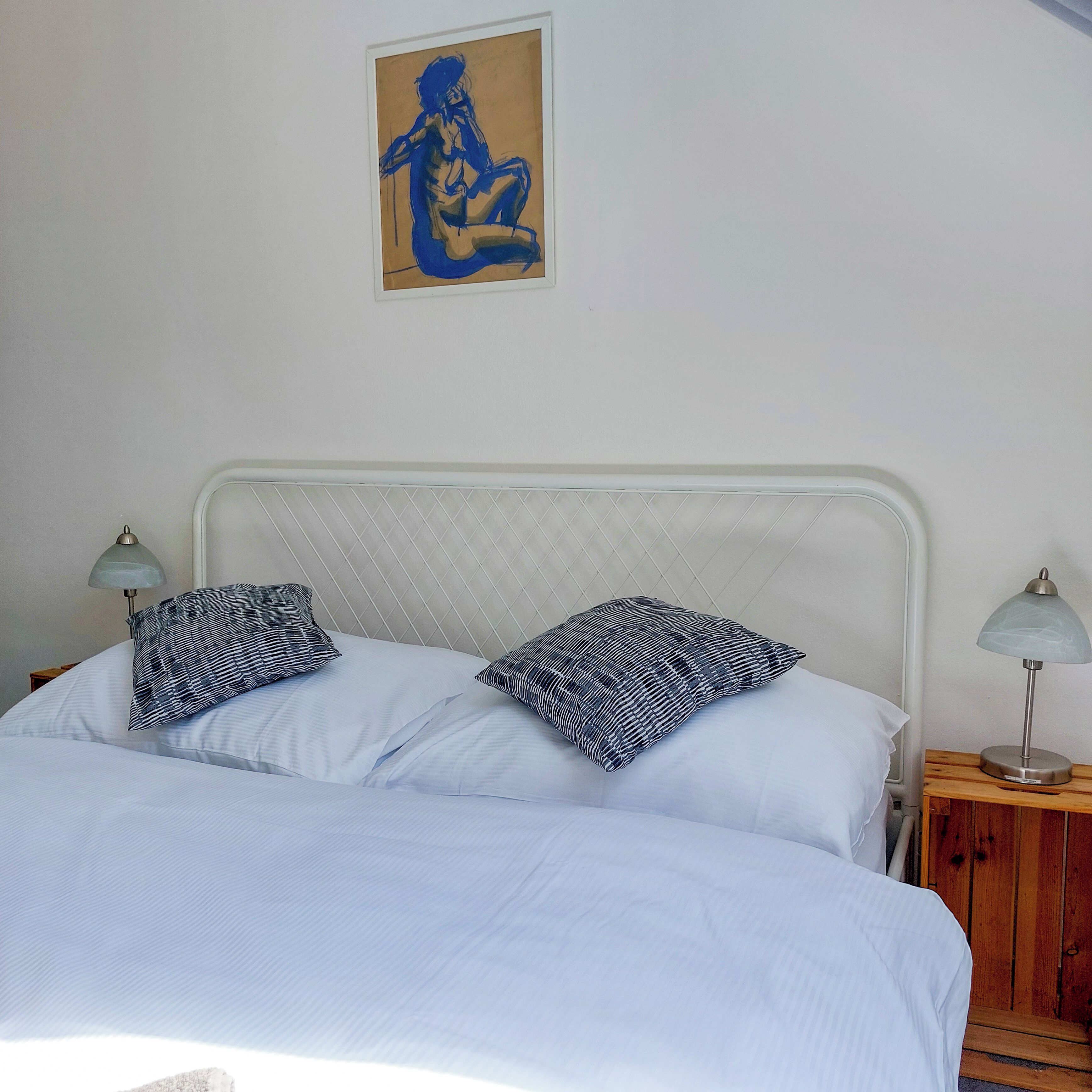 Una cama con ropa de cama blanca y dos almohadones a rayas, una mesita de noche con lámpara y un cuadro en la pared.